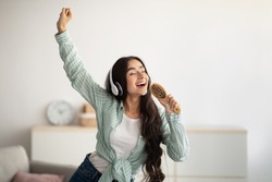 Happy Indian lady with headphones dancing to music and singing song, using hairbrush as mic at home. Young woman moving to her favorite soundtrack, pretending to be rock star