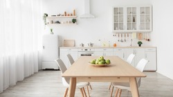 Contemporary minimalist interior of kitchen and dining room. White furniture with utensils and dinner table with chairs in Scandinavian style. Modern light design
