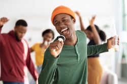Positive african american young man with bottle of beer singing karaoke while having home party with friends, holding microphone and singing songs, selective focus, blurred background