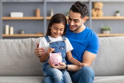 Financial Education For Children Concept. Portrait of smiling little girl putting coin in pink piggy bank, sitting on dad's lap on the couch at home, man teaching his daughter to invest