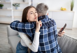 Beautiful mature woman hugging her husband, showing HUSH gesture, having secret affair, texting her lover on smartphone. Adultery, relationship issue, marriage cheating, hidden love concept