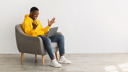 Excited African American Man With Laptop Shouting Celebrating Great Online News Sitting In Chair Over Gray Wall Indoor, Side View. Successful Freelance Career Concept. Panorama, Copy Space