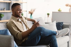 Great Mobile Application. Happy African American Man Using Phone Texting And Browsing Internet Sitting On Sofa At Home, Side View. Black Male Networking In Social Media Using App On Smartphone