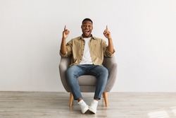 Excited African American Millennial Man Pointing Fingers Up Sitting In Chair Over Gray Wall Background, Looking At Camera. Check This Our, I Have Great Idea Concept. Front View
