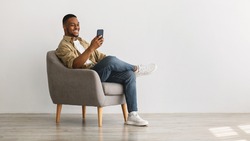 Cheerful African American Guy Using Mobile Phone With New Application Sitting In Armchair Over Gray Wall Background. Gadgets And Mobile Communication Concept. Panorama, Empty Space For Text