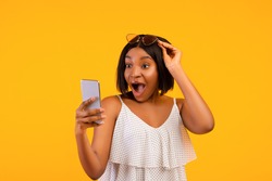 Shocked black lady in summer outfit looking at smartphone in excitement, lifting her sunglasses on orange studio background. Surprised African American woman learning about online summertime sale