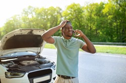 Unhappy black man standing near his broken car with open hood, experiencing engine failure, having mobile phone conversation, calling breakdown road service on smartphone