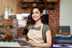 Opening small business. Happy arab woman in apron near bar counter holding digital tablet and looking at camera, waiting for clients in modern loft cafe