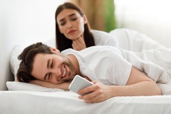 Online Affair. Wife Spying While Cheating Husband Texting On Cellphone Having Flirty Chat Conversation With Lover Lying In Bed At Home. Jealous Woman Checking Her Boyfriend's Phone. Selective Focus