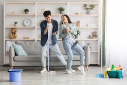 Funny asian loving couple singing songs while cleaning apartment, using broom and mop as microphones, cheerful beautiful young man and woman imitating rock stars while house-keeping, copy space