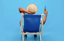 Back view of young black guy sitting in lounge chair with bottle of beer, enjoying summer vacation, sipping alcoholic beverage, sunbathing on blue studio background, full length