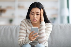 Bad News. Upset Asian Female Holding Smartphone, Looking At Mobile Phone Screen With Worry While Sitting On Couch At Home, Anxious Korean Woman Touching Head While Reading Unplesant Message, Closeup