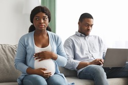Lonely pregnant african american woman sitting by her ignoring husband using laptop, chatting with his friends. Sad expecting black lady looking for attention from her man, living room interior