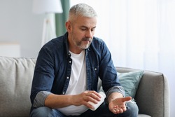 Senior man taking pills while sitting on couch at home, holding white jar with treatment, copy space. Grey-haired elderly man using supplements or vitamins. Healthy lifestyle in senior age concept