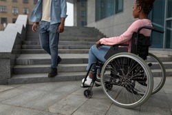 Young black disabled woman suffering from lack of wheelchair friendly facilities, looking at man going down stairs, cannot get home without ramp. Handicapped people problems concept