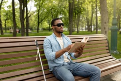 Young black visually impaired man sitting on bench in city park, reading Braille book outdoors. Millennial blind guy in dark glasses studying or enjoying entertainment literature, outside