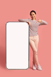Joyful Asian Lady Leaning And Pointing At Big Smartphone With Blank White Screen, Demonstrating Copy Space For Your App Or Website Design, Standing On Pink Studio Background, Mockup Image