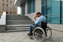 Disability and accessible environment problems. Upset handicapped black man in wheelchair in front of stairs without ramp, having no possibility to enter building, copy space