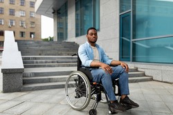 Accessible facilities for disabled people. Portrait of unhappy black impaired man in wheelchair next to siatrs without ramp, feeling stressed, having difficulty in entering building, free space