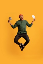 Big Win. Portrait Of Excited Black Guy Jumping In Air With Dollar Cash In Hands, Young African American Man Holding Lots Of Money And Screaming With Joy Over Yellow Studio Background, Copy Space