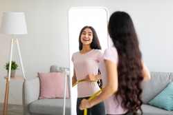 Attractive young Indian woman measuring her waist with tape near mirror indoors, copy space. Young Asian lady happy with results of slimming diet or liposuction, being successful in weight loss