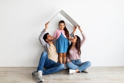 Housing for young family concept. Young eastern father, mother and daughter under symbolic roof dreaming of new home, sitting on floor over light wall