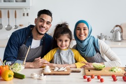 Portrait Of Happy Young Middle Eastern Family Cooking Together In Kitchen, Cheerful Arabic Parents And Their Little Daughter In Aprons Sitting At Table While Preparing Tasty Food Together