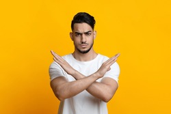 Serious arab guy showing stop or no gesture over yellow studio background, copy space. Annoyed middle-eastern young man in white t-shirt crossing hands, dislike or deny something