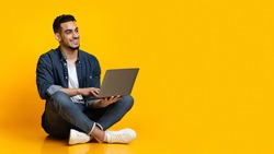 Happy arab young man sitting on floor, using new modern laptop, typing on keyboard and smiling over yellow studio background, looking at copy space for text or advertisement, panorama