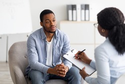 Unhappy young black man having session with professional psychologist at mental health clinic. Psychotherapist taking notes during conversation with depressed male patient