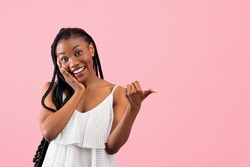 Joyful black lady pointing at blank space, touching her face in excitement over pink studio background. Charming African American woman advertising your product, promoting sale or discount