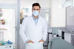Young doctor in medical face mask standing at dental clinic, copy space. Handsome male dentist greeting patients or inviting to visit brand new modern dental clinic, healthcare and pandemic concept