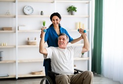Post-stroke rehabilitation for disabled people. Millennial physiotherapist helping elderly male patient in wheelchair to make exercises at home. Impaired senior man working out with dumbbells