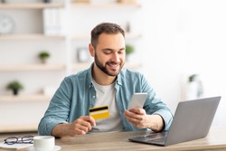Happy Caucasian man buying things online, using smartphone, laptop and credit card, enjoying shopping in internet. Positive young guy purchasing goods on web, making remote payment