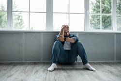 Man feeling depressed and desperate, crying alone at home, suffering, emotional pain, unhappy, mental health issues. Sad young bearded guy sits on floor near large window and holds his head with hand
