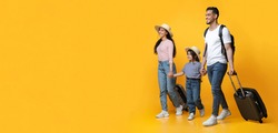 Happy cheerful middle eastern family of three with suitcases going on summer vacation together, joyful arab parents and their little daughter walking with luggage on yellow studio background, panorama