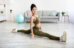 Sporty Indian woman sitting in splits, doing legs stretch, improving flexibility at home, full length. Athletic Eastern lady keeping fit and healthy during covid quarantine, copy space