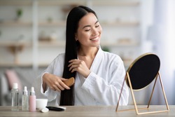 Cheerful young asian lady in bathrobe sitting in bedroom, combing her beautiful silky hair with wooden brush, looking at mirror and smiling, copy space. Hair care products concept