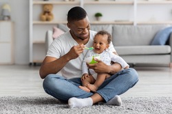 Happy Young Black Dad Spoon Feeding His Cute Adorable Baby Son At Home, Loving African American Father Taking Care About Little Male Infant Child Enjoying Paternity Leave Time, Free Space