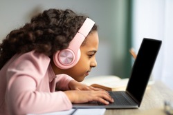 Youth and technology concept. Focused black schoolgirl using laptop computer, sitting at table too close to pc, side view. Little girl with poor eyesight having near vision problem