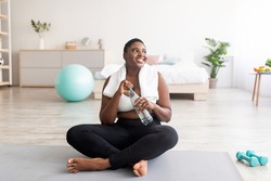 Plump black lady in sportswear sitting on sports mat with bottle of clear water at home, full length. Curvy African American woman keeping hydrated during domestic training indoors