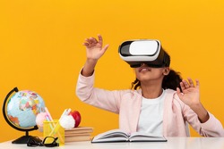New Experience In Education And Knowledge. Black teenager using modern VR glasses for studying, gesturing on orange yellow studio background, sitting at desk with globe and books, free copy space