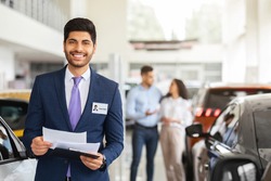 Charismatic arab man in suit sales assistant posing over auto showroom, holding chart with papers and smiling at camera, standing over various cars and customers choosing new automobile, copy space