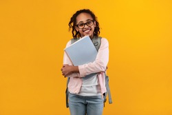 Education Concept. Portrait of positive smiling African American girl wearing glasses and backpack holding textbooks, posing and looking at camera isolated on yellow studio background