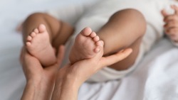 Maternal Love Concept. Closeup of young mom holding feet of her cute little African American baby in hands. Child wearing bodysuit lying on the white blanket sheets at home, selective focus on legs