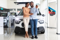Beautiful african american couple bought car in dealership center, happy black spouses posing with keys and embracing, celebrating purchasing new family vehicle in automobile showroom, free space