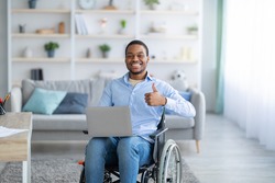 Portrait of happy handicapped black guy with laptop showing thumb up gesture, smiling at camera indoors. Paraplegic young man in wheelchair recommending online job during covid pandemic