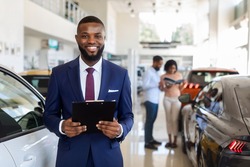 Portrait Of Handsome Black Car Salesman In Suit Posing At Workplace In Auto Showroom, Young Dealership Center Manager With Clipboard In Hands Helping Spouses To Purchase New Automobile, Free Space
