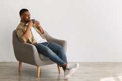 Contented Black Man Enjoying Morning Coffee Sitting In Comfortable Chair On Gray Background Indoors, Looking Aside At Free Space. Weekend Relaxation Concept. Side View