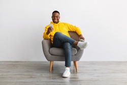 Great App. Contented Black Man Using Smartphone Advertising New Mobile Application, Texting Online Sitting In Armchair Near Gray Wall Indoors. Modern Mobile Communication, People And Gadgets